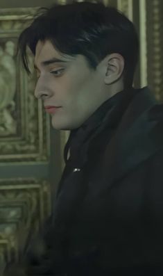 a man with black hair and piercings looks off into the distance while standing in front of an ornate wall