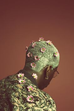 a man with flowers on his head and glasses in front of a brown background wearing a green sweater