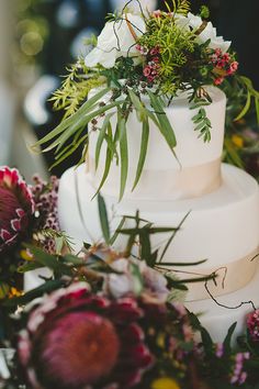 a white wedding cake with flowers and greenery on the top is surrounded by other floral arrangements