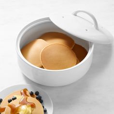 two plates with pancakes and blueberries on them next to a white container filled with pancakes