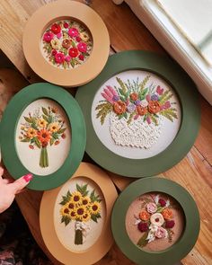 four hand embroidered flowers are displayed in green frames on a wooden floor next to a woman's finger