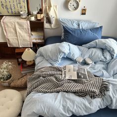 an unmade bed with blue sheets and pillows in a small room filled with books