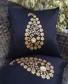 two black pillows with gold embroidered designs on them sitting next to a wicker chair