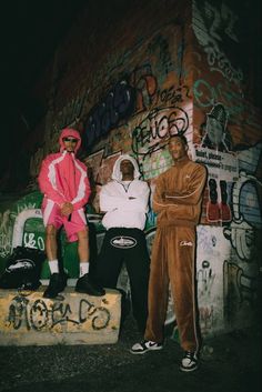 two men sitting on top of a cement block in front of graffiti covered wall with one man wearing a pink and white hoodie