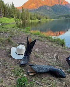 two cowboy boots and hats sitting on the ground next to a lake