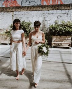 two women in white dresses holding hands and walking down a sidewalk with graffiti on the wall behind them