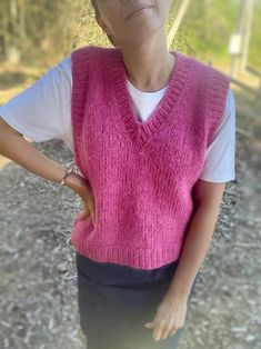 a woman wearing a pink sweater vest standing on a dirt road with her hands in her pockets