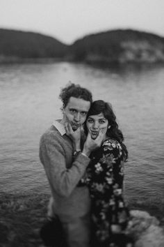 a man and woman standing next to each other in the water with mountains in the background