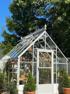 a white greenhouse with potted plants in the corner and trees around it on a sunny day