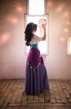 a woman in a long purple dress looking out a window at the sun coming through