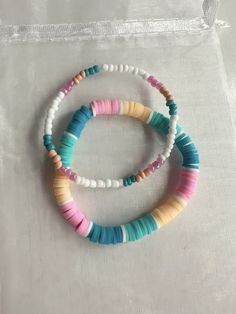 two bracelets with different colored beads are on a white cloth covered bag, and one is in the shape of a heart