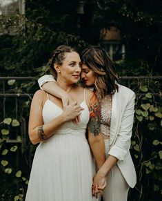 two women are hugging each other in front of some trees and bushes, one is wearing a white dress