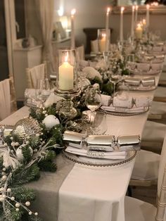 the table is set with silver and white dishes, place settings, candles and greenery