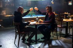 two men sitting at a table with food and drinks in front of them, one holding up his hand