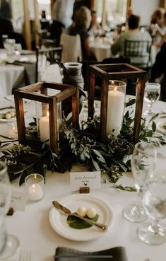 two candles are sitting on top of a white table cloth with greenery and silverware