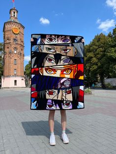 a woman holding up a piece of art with multiple faces on it in front of a clock tower