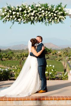 a bride and groom kissing under an arch with white flowers on the altar at their wedding