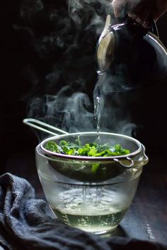 someone pouring water into a glass bowl filled with green vegetables and steam rising from it