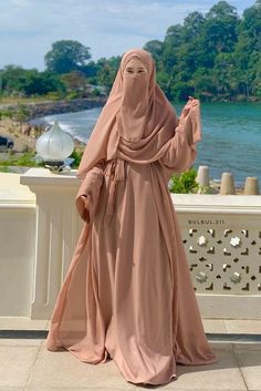 a woman in a hijab is standing on a ledge near the water and looking off into the distance