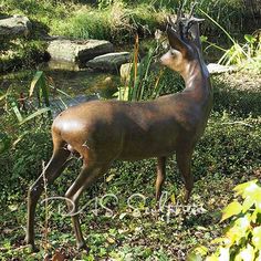 a statue of a deer standing in the grass next to a stream and trees with green foliage