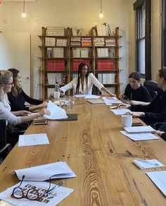 a group of people sitting around a wooden table with papers on it and one person standing in front of them