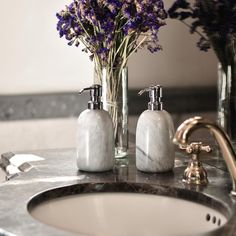 two vases filled with purple flowers sitting on top of a bathroom counter next to a sink