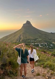 two women walking down a path towards the ocean with mountains in the background at sunset