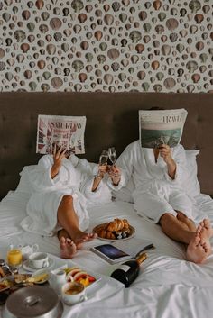 two people sitting on a bed with food and drinks in front of them reading magazines