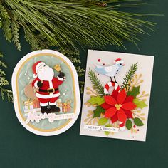 two christmas cards sitting on top of a table next to a pine branch and evergreen