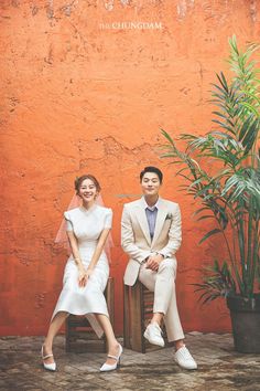 a man and woman sitting next to each other in front of a wall with plants