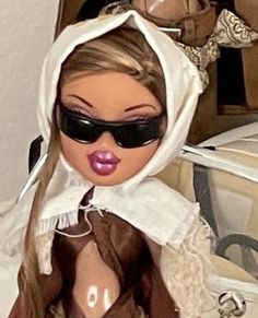 a close up of a doll wearing sunglasses and a hood over her head with a suitcase in the background