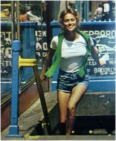 a woman in short shorts and a green vest is standing on the sidewalk with her legs crossed