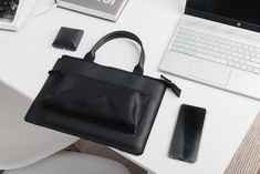 a black purse sitting on top of a white table next to a laptop and cell phone