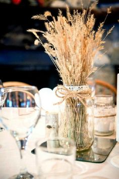 the table is set with wine glasses and vases filled with dried grass in them