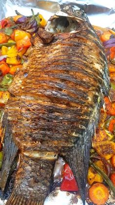 a large fish is laying on some tin foil with other food items around it, including carrots and peppers