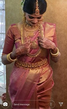 a woman in a pink and gold sari with jewelry on her neck, standing next to a window