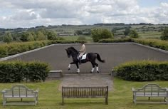 a woman riding on the back of a black horse across a lush green field next to park benches