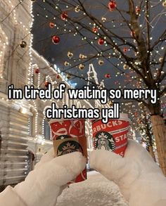 someone holding up two starbucks cups in front of a christmas tree with lights on it