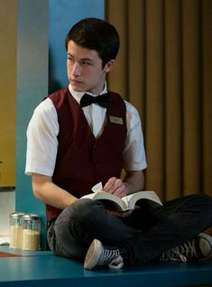 a young man sitting on the floor reading a book while wearing a vest and bow tie