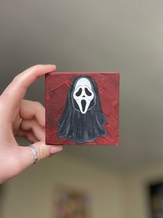 a person holding up a small square painting with a ghost on it's face