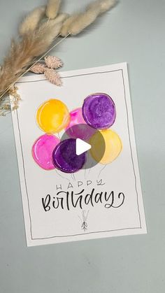 a happy birthday card with balloons and feathers on the table next to some dried flowers