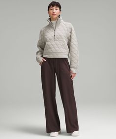 a woman standing in front of a gray background wearing brown pants and a white sweater