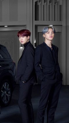 two men standing next to each other in front of a black car with red hair