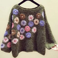 a green sweater with flowers on it is hanging from a hanger and someone's hand is holding the top