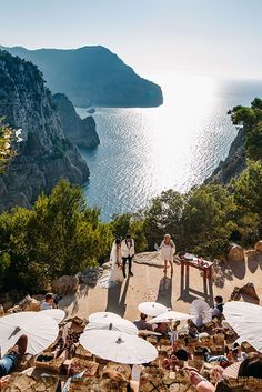 people are sitting at tables on the cliff by the water with umbrellas in front of them