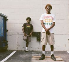 two young men standing next to each other in front of a white wall and blue trash can