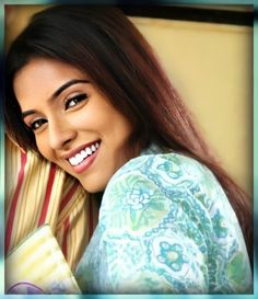 a woman with long brown hair smiling and wearing a colorful shirt, leaning against a yellow wall