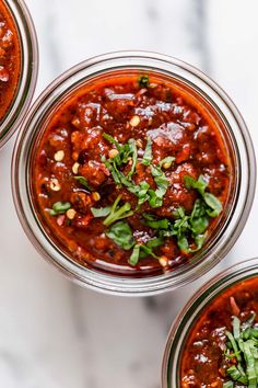 three jars filled with chili sauce and garnished with fresh herbs on top, sitting on a marble surface