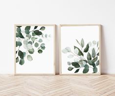 two framed art prints with green leaves on white wall above chevron wood flooring