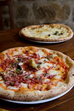 two pizzas sitting on top of a wooden table next to each other in front of a stone wall